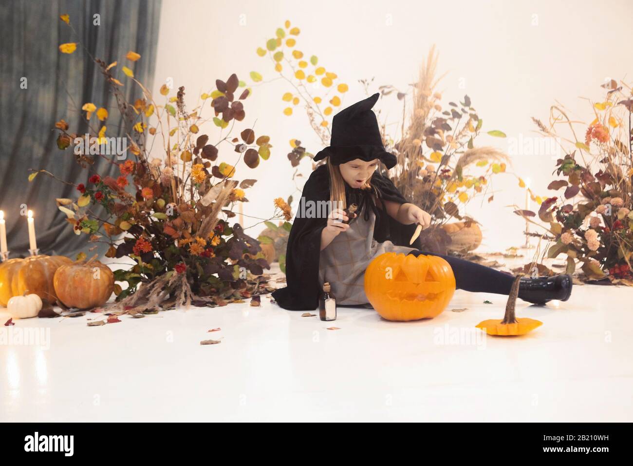 Little cute girl with long hair standing on scene in black dress and hat with autumn decorations on background during Halloween Stock Photo