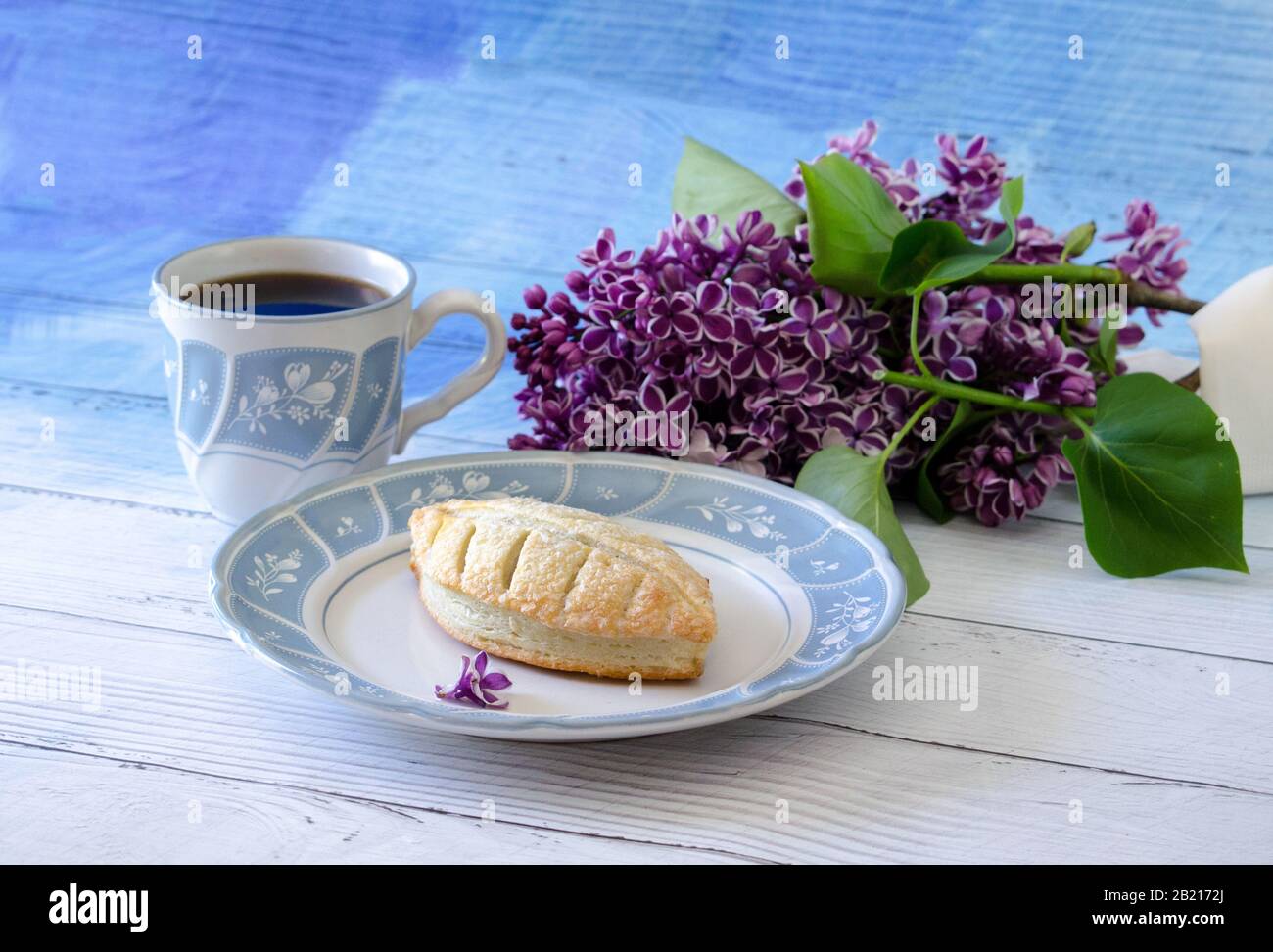 A puff pastry is filled with chocolate ad served with a cup of hot coffee and a beautiful sprig of lilac flowers Stock Photo