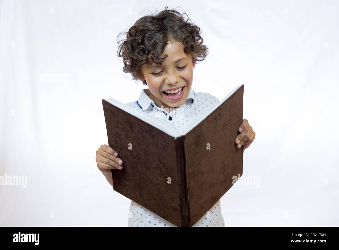 Smiling boy between 8 and 9 years old holding a book while reading it and standing against white background Stock Photo