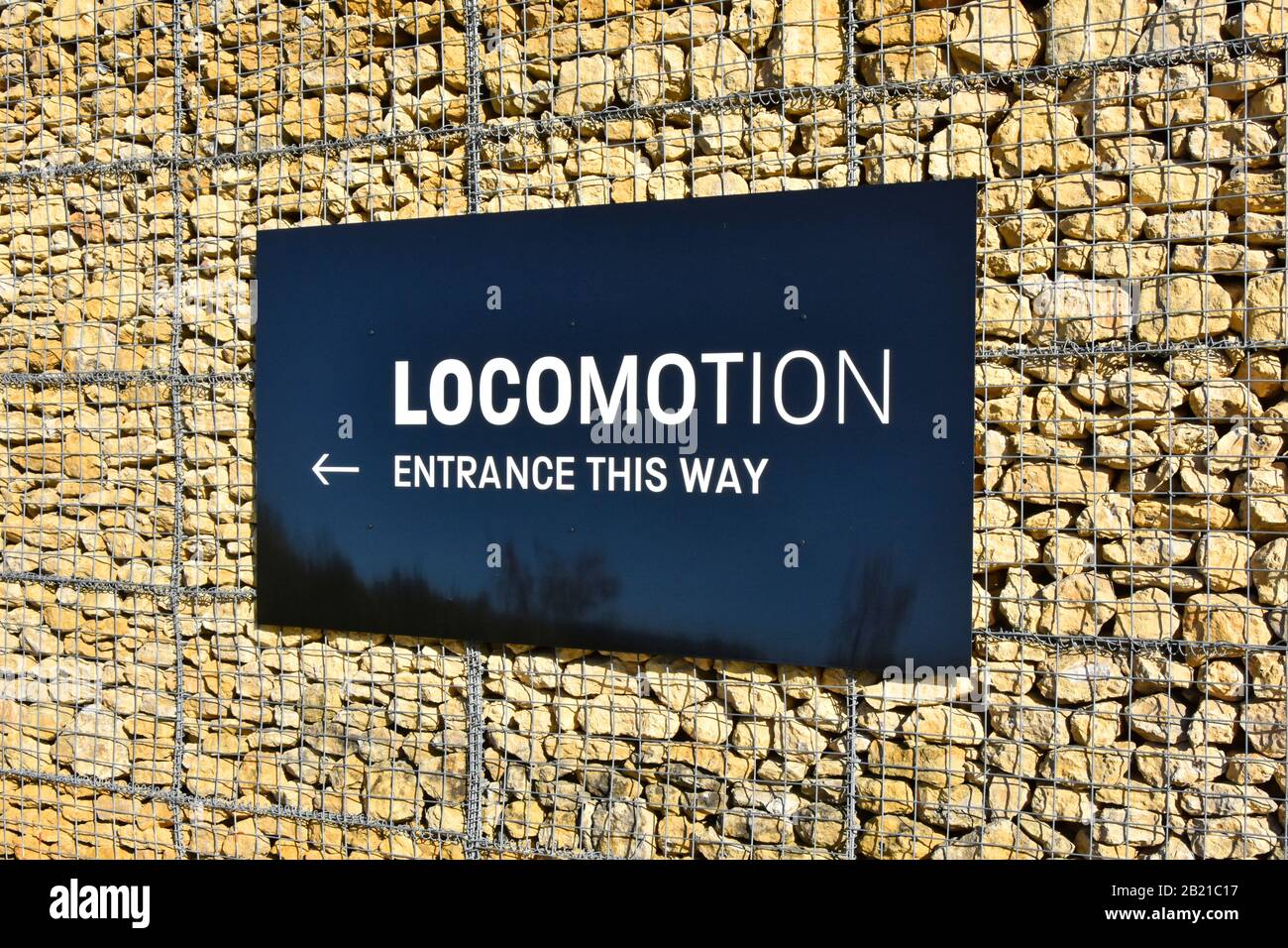 Sign for Locomotion  Museum entrance fixed on Gabion wire cage of broken quarry stone forming external battered building wall Shildon County Durham UK Stock Photo