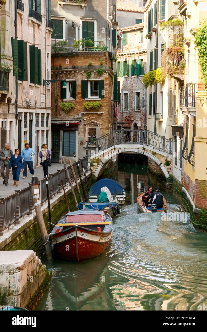 Motorboat going quickly through the canal. People walking along the walkway and buildings. Residential area of Venice, Italy. NMR Stock Photo