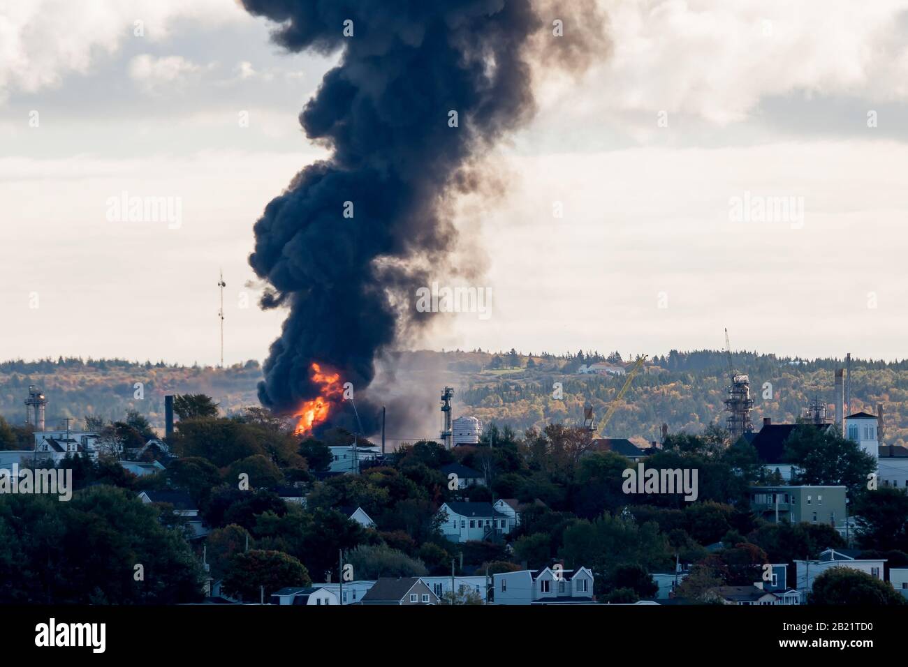 Large fire at an oil refinery seen from a distance. Only parts of the refinery visible behind the trees. Thick black smoke rises from the flames. Stock Photo