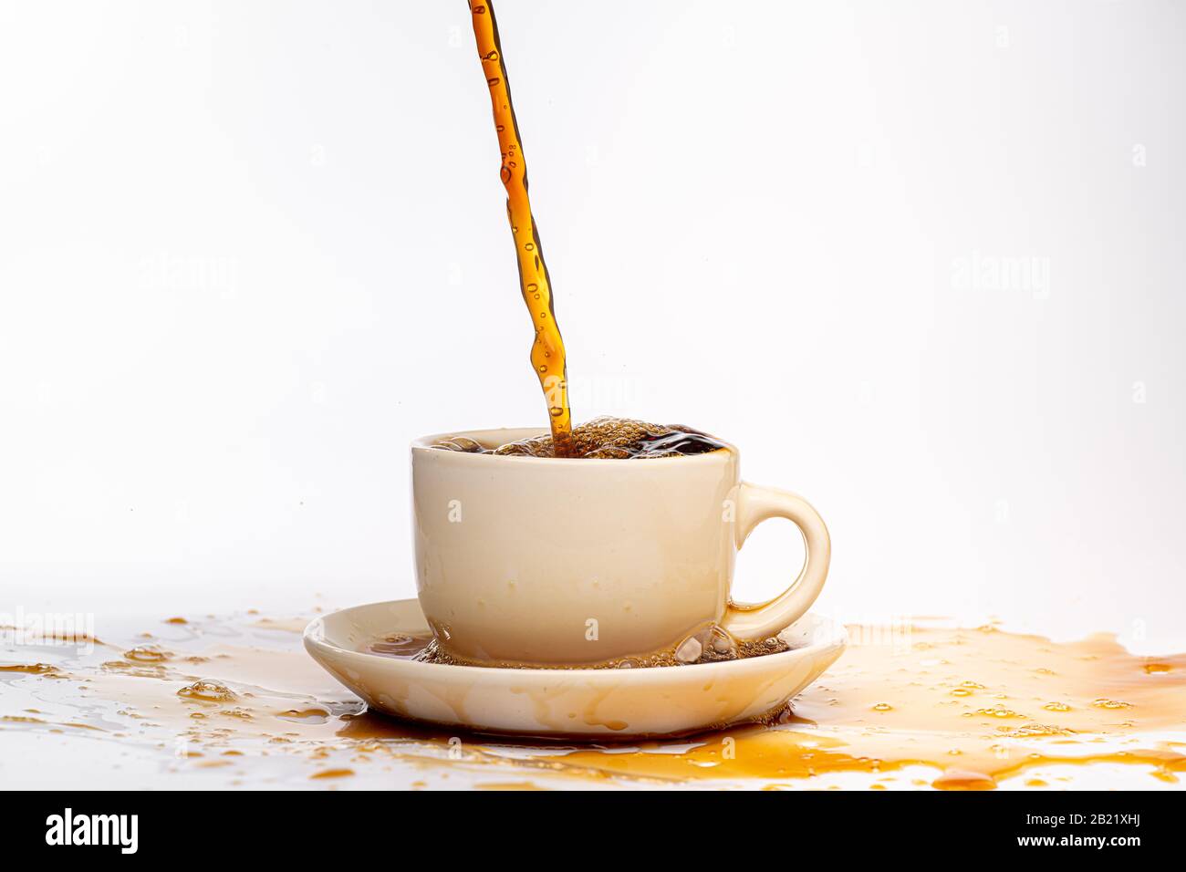 Coffee pouring into a white cup isolated against a plain white background, splashing in all directions creating a mess on the white surface Stock Photo