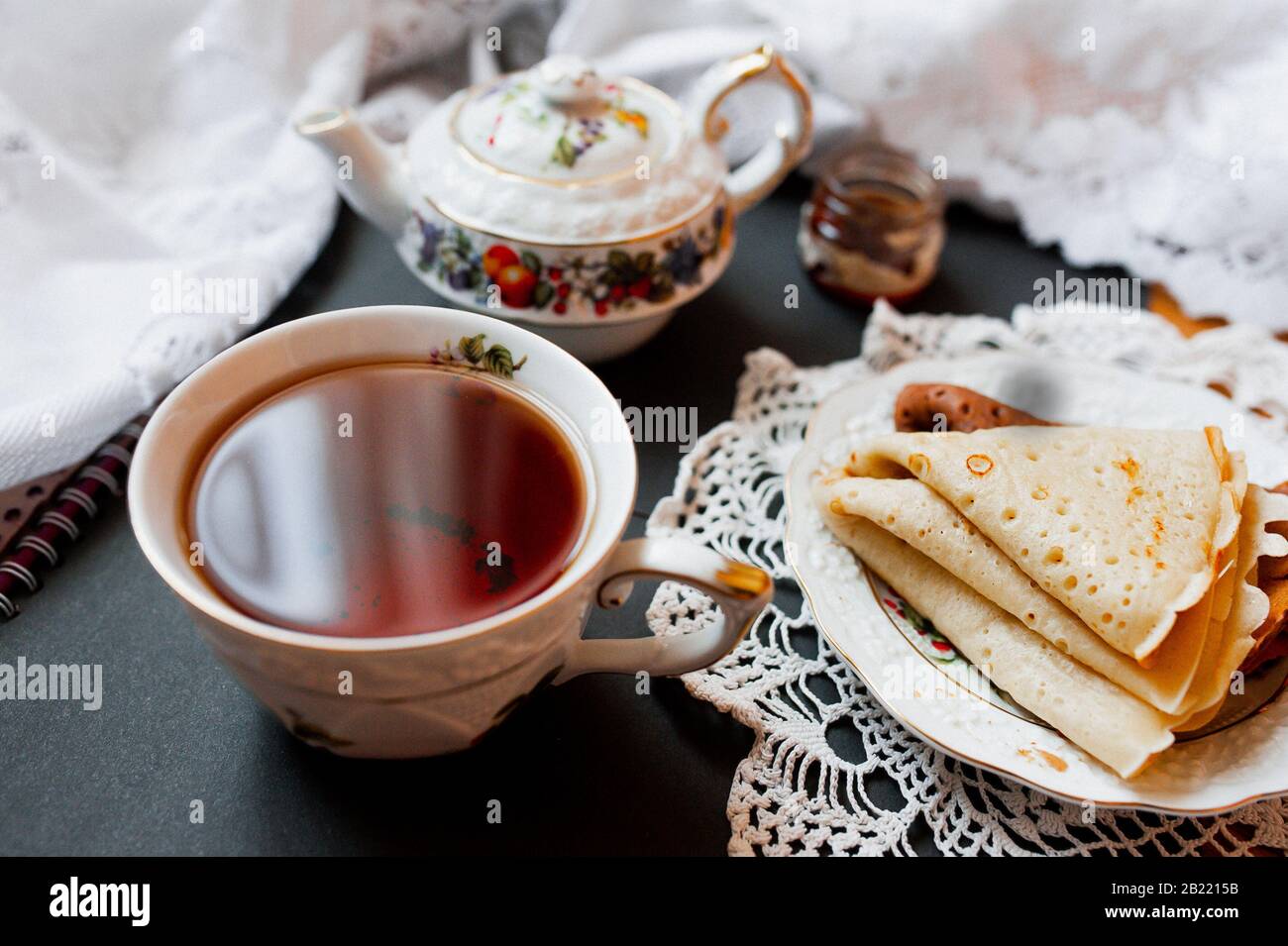 A beautiful version of Breakfast with a white porcelain Cup and a painted teapot in the background, ready-made pancakes on a saucer. Table setting for Stock Photo