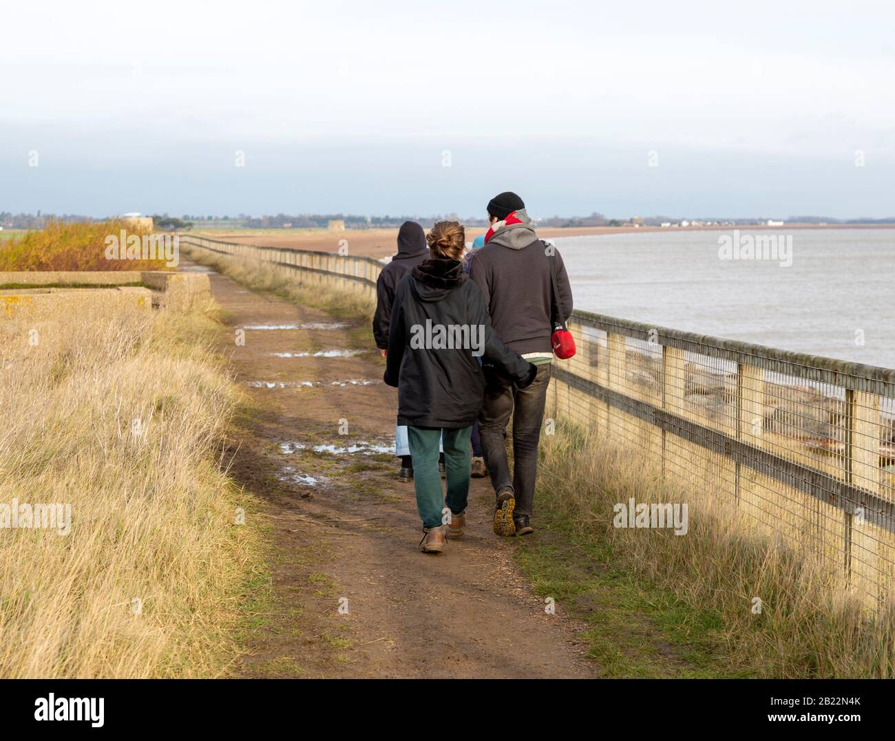 View from behind of people wearing winter coats walking on path by North Sea coast, Bawdsey, Suffolk, England, UK Stock Photo