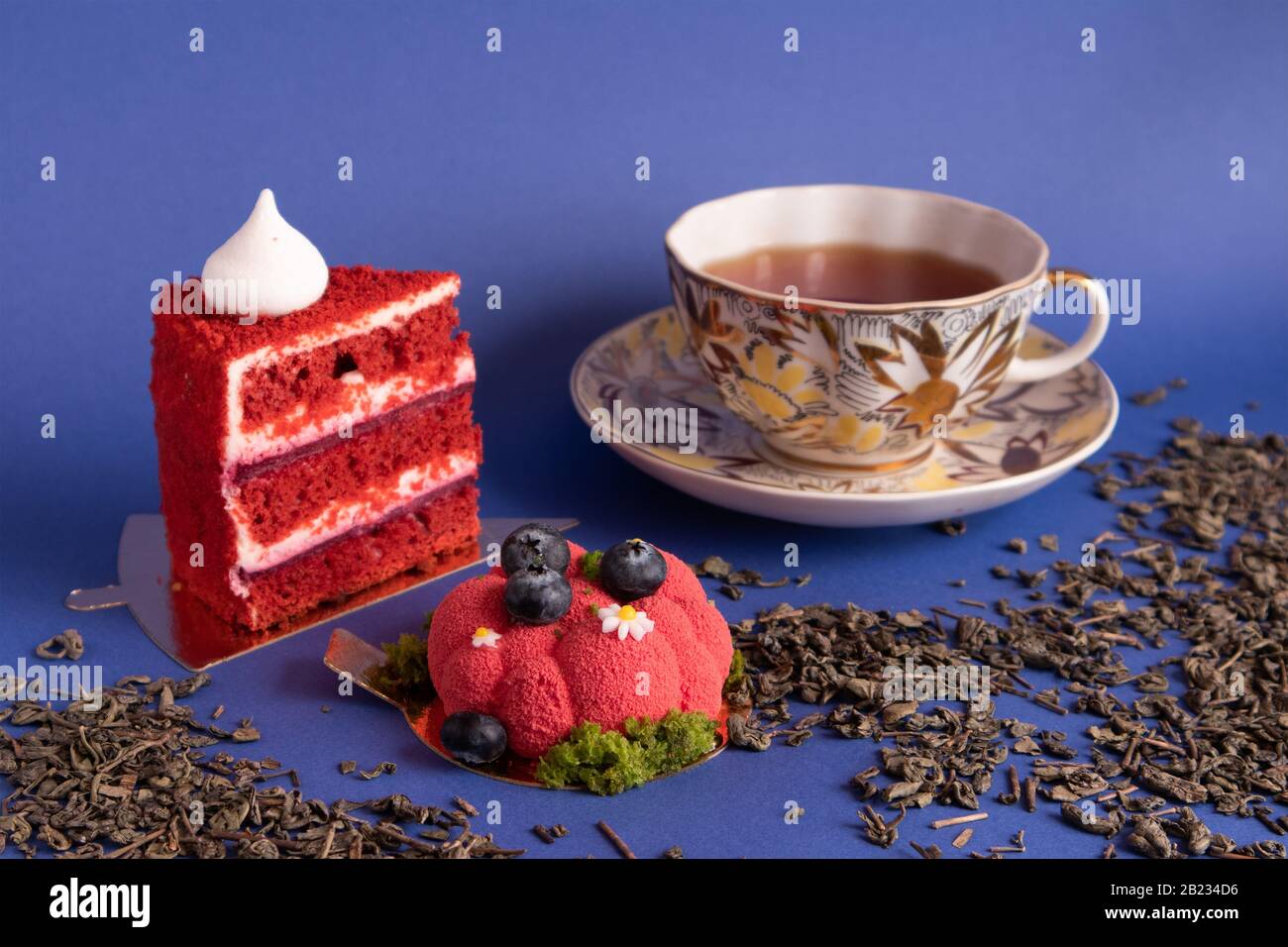 Two appetizing delicious red cakes and a cup of tea on a violet background with scattered dry green tea Stock Photo