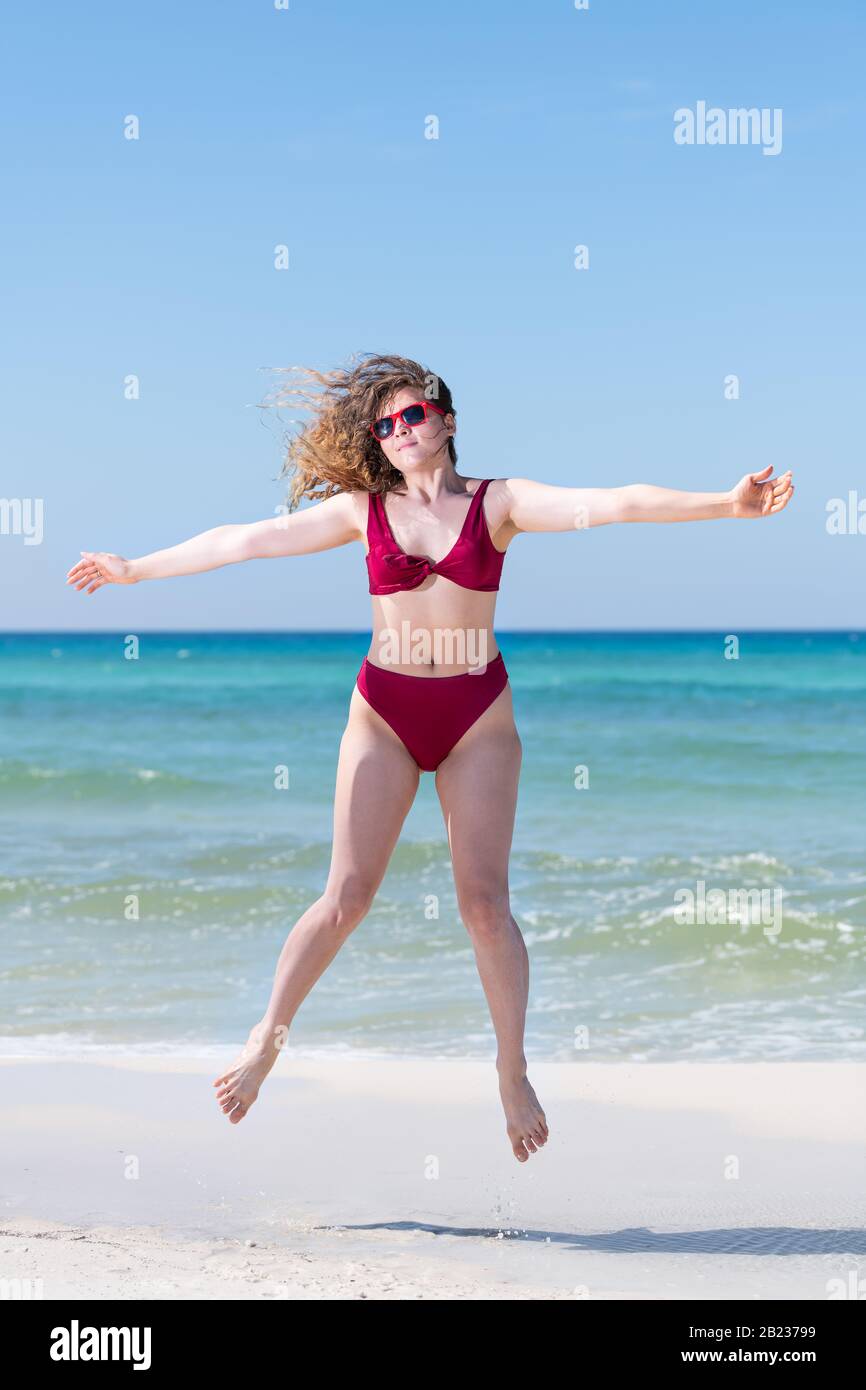 Young girl in red bikini bathing suit sunglasses jumping up with ocean