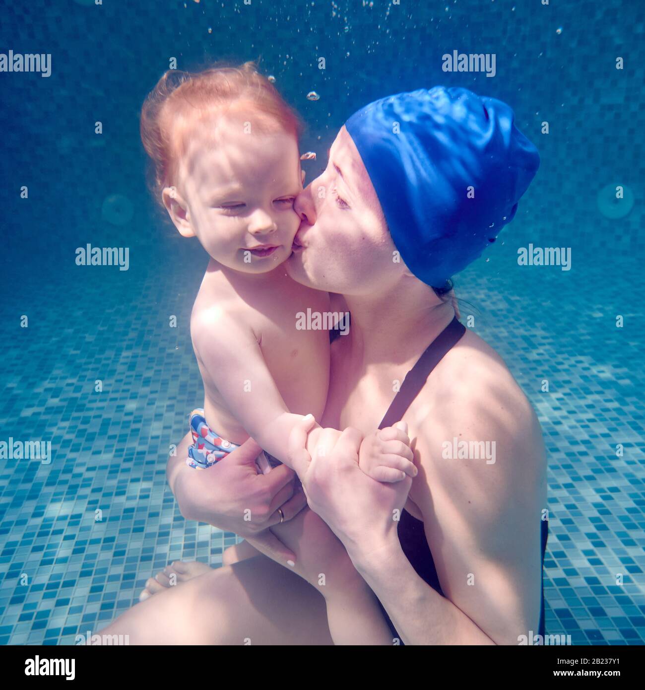 Young woman in swim cap holding cute child in arms and placing kiss on baby cheek. Charming lady sitting on swimming pool floor underwater and kissing son. Concept of swimming, family and mother love. Stock Photo