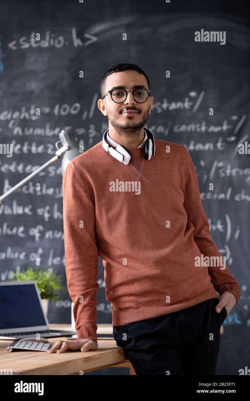 Young confident it-manager in casualwear and eyeglasses standing by desk with laptop on background of blackboard with formula Stock Photo