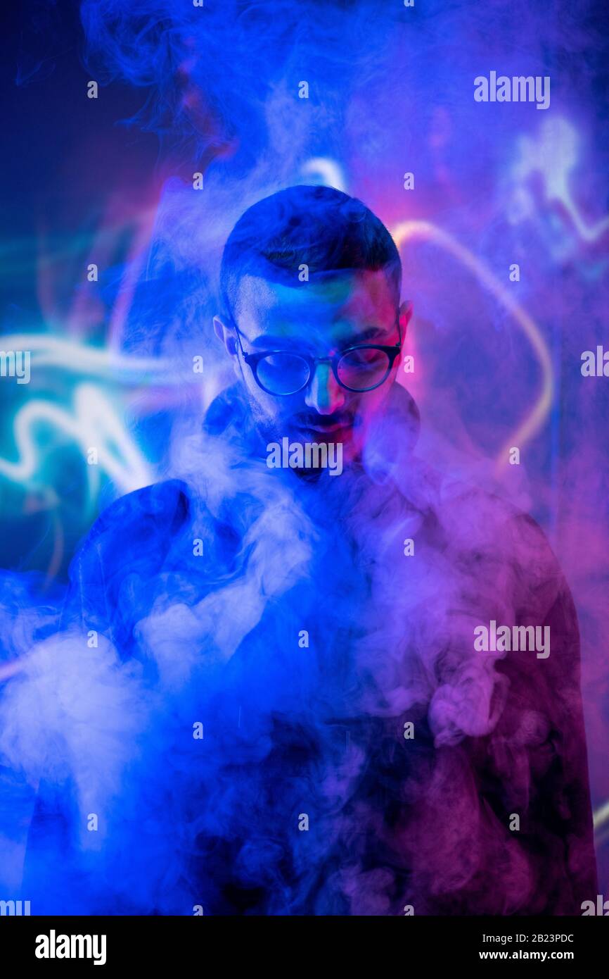 Head of young man in eyeglasses standing in isolation in front of camera among smoke and blue neon light Stock Photo
