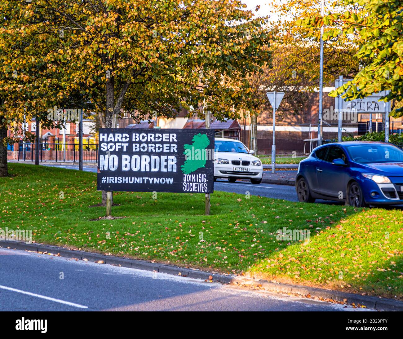 A sign reading "Hard Border, Soft Border, No Border – #IrishUnityNow, Join 1916 Societies," with a green map of Ireland, stands beside a road in the Bogside area of Londonderry, County Derry, Northern Ireland; "RIRA" graffiti is visible on a wall in the background. The 1916 Societies are an Irish republican network campaigning for a united Ireland. Stock Photo