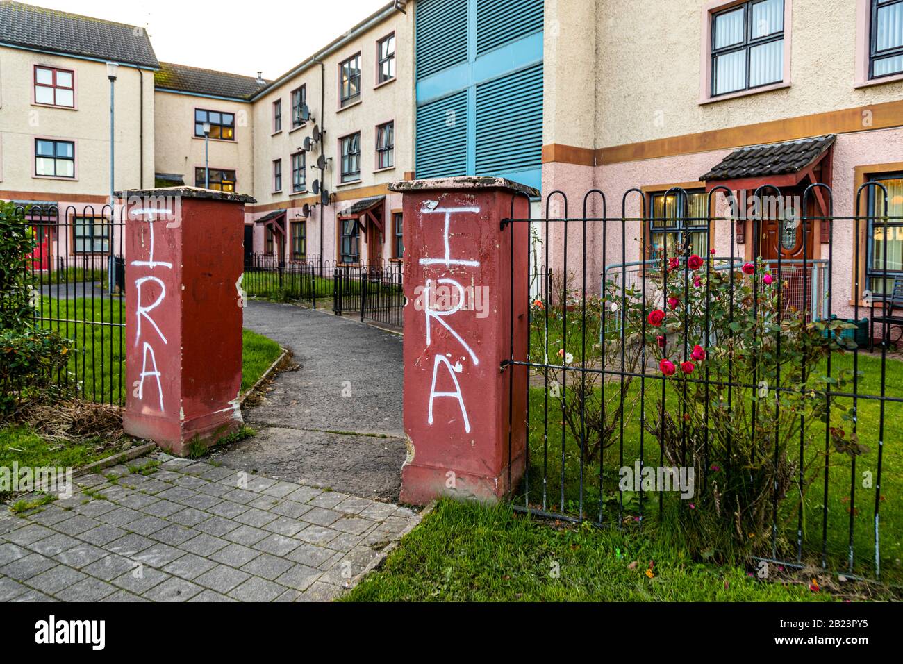 "IRA" graffiti has been spray-painted on two gate pillars in front of a housing block in the Bogside area of Londonderry, County Derry, Northern Ireland; roses bloom in the front garden behind them. Stock Photo