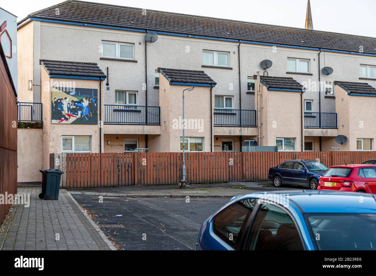 A smaller mural decorates the gable end of a modern terraced house in the Bogside district of Londonderry, County Derry, Northern Ireland. It shows a turbulent street scene with several figures and a red-marked shape lying on the ground – imagery from the era of the Northern Ireland conflict ("the Troubles", 1968–1998). Parked cars and a wooden fence line the front of the terrace. Stock Photo