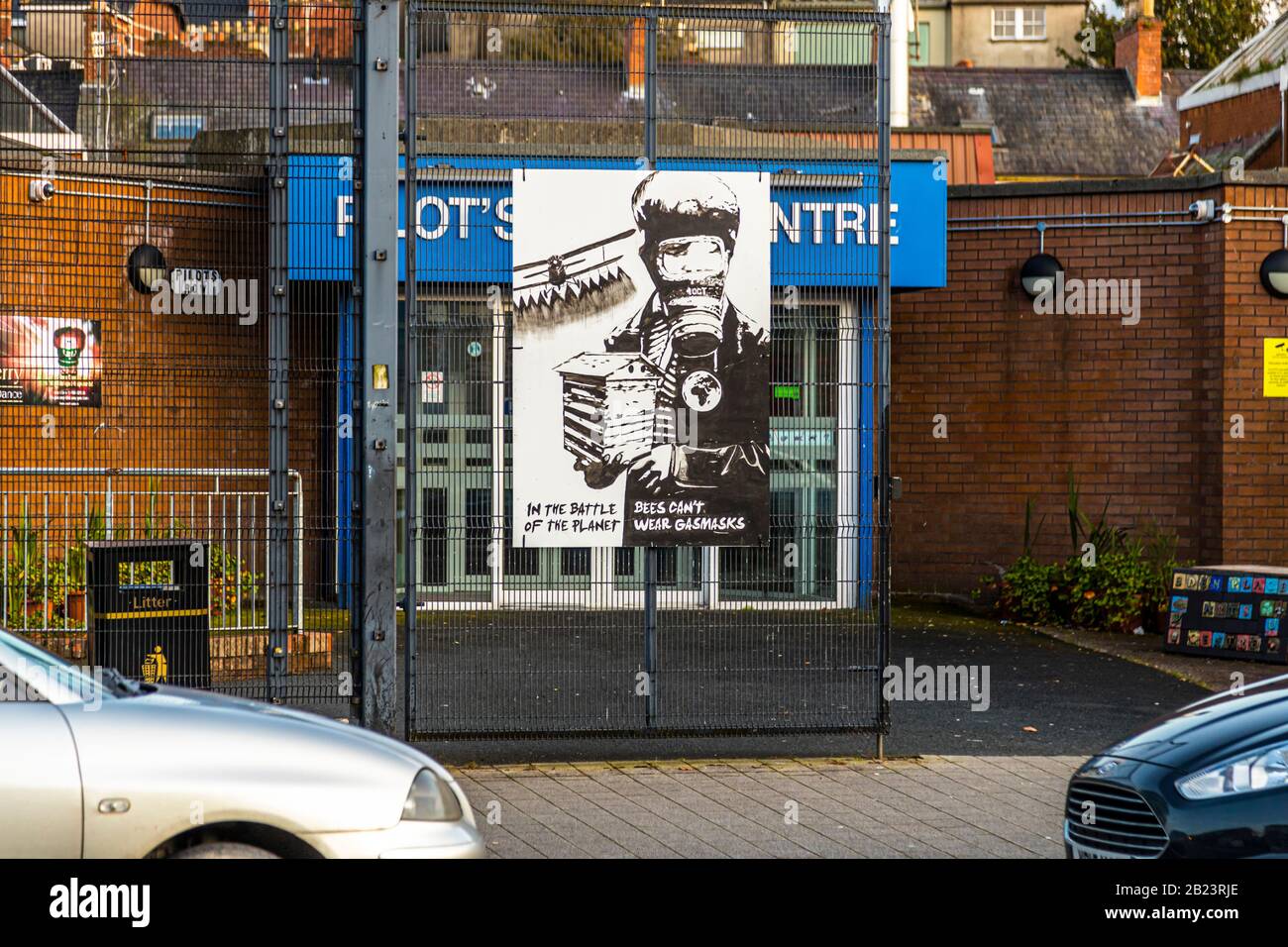 A poster showing a figure in a gas mask carrying a beehive hangs on the fence in front of a building in the Bogside area of Londonderry, County Derry, Northern Ireland. The text "In the Battle of the Planet, Bees Can't Wear Gasmasks" links environmental awareness to the area's local gas-mask imagery from the era of the Northern Ireland conflict. Parked cars line the street in front. Stock Photo