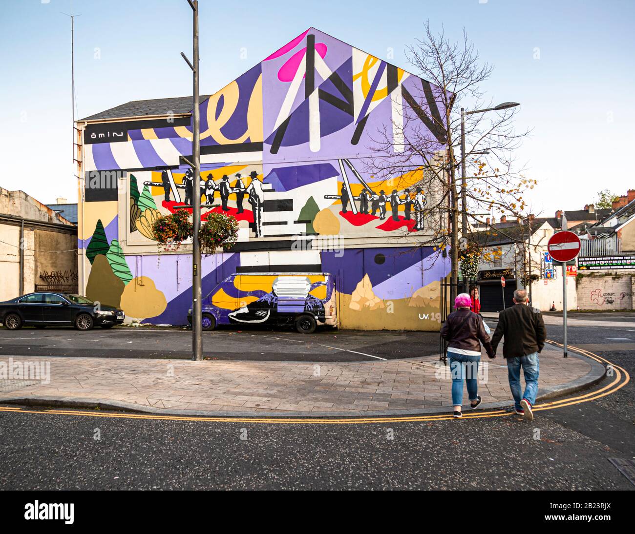 A colourful abstract mural with geometric shapes and a stylised vehicle scene decorates a building on Rossville Street in the Bogside area of Londonderry, County Derry, Northern Ireland – signed "UV19", contemporary street art unlike the surrounding political murals, with no apparent link to the Northern Ireland conflict. A couple walks hand in hand past the building. Stock Photo