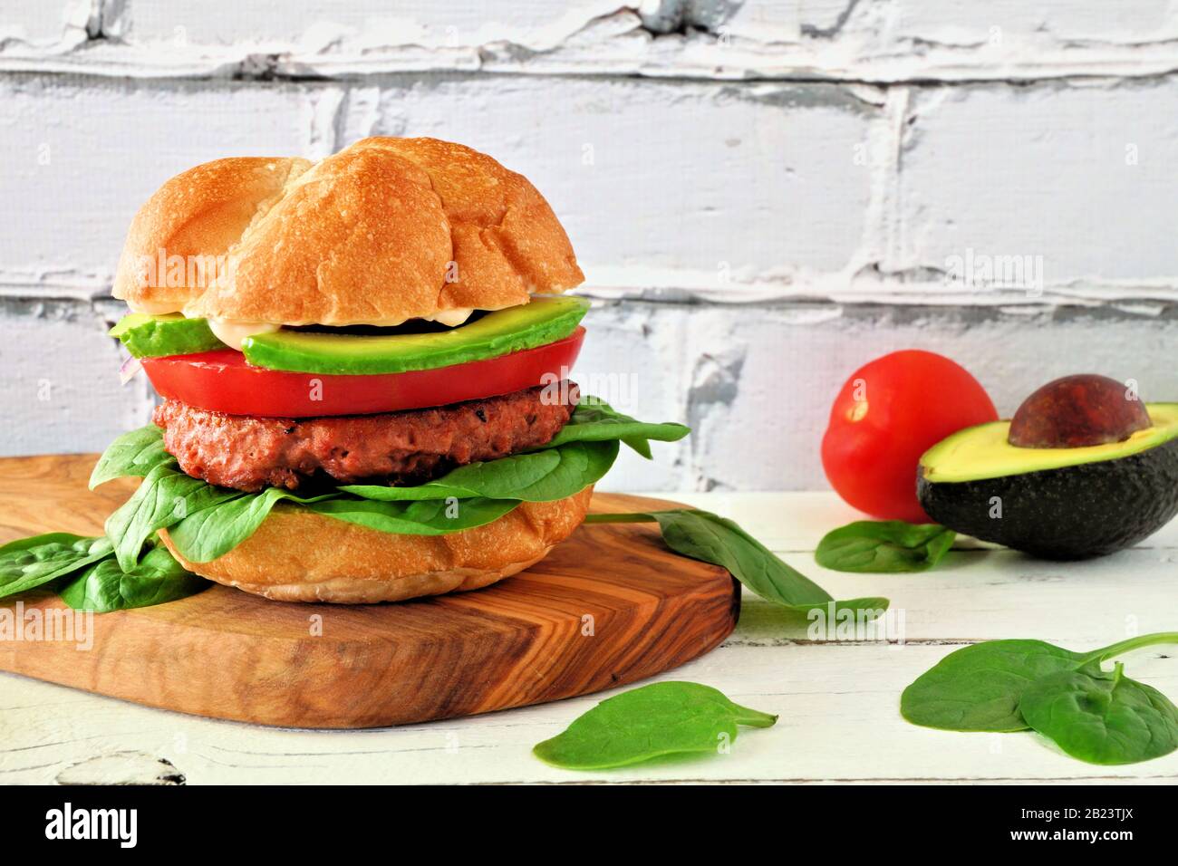Plant based meatless burger with avocado, tomato and spinach on a wood serving board against a white brick background Stock Photo