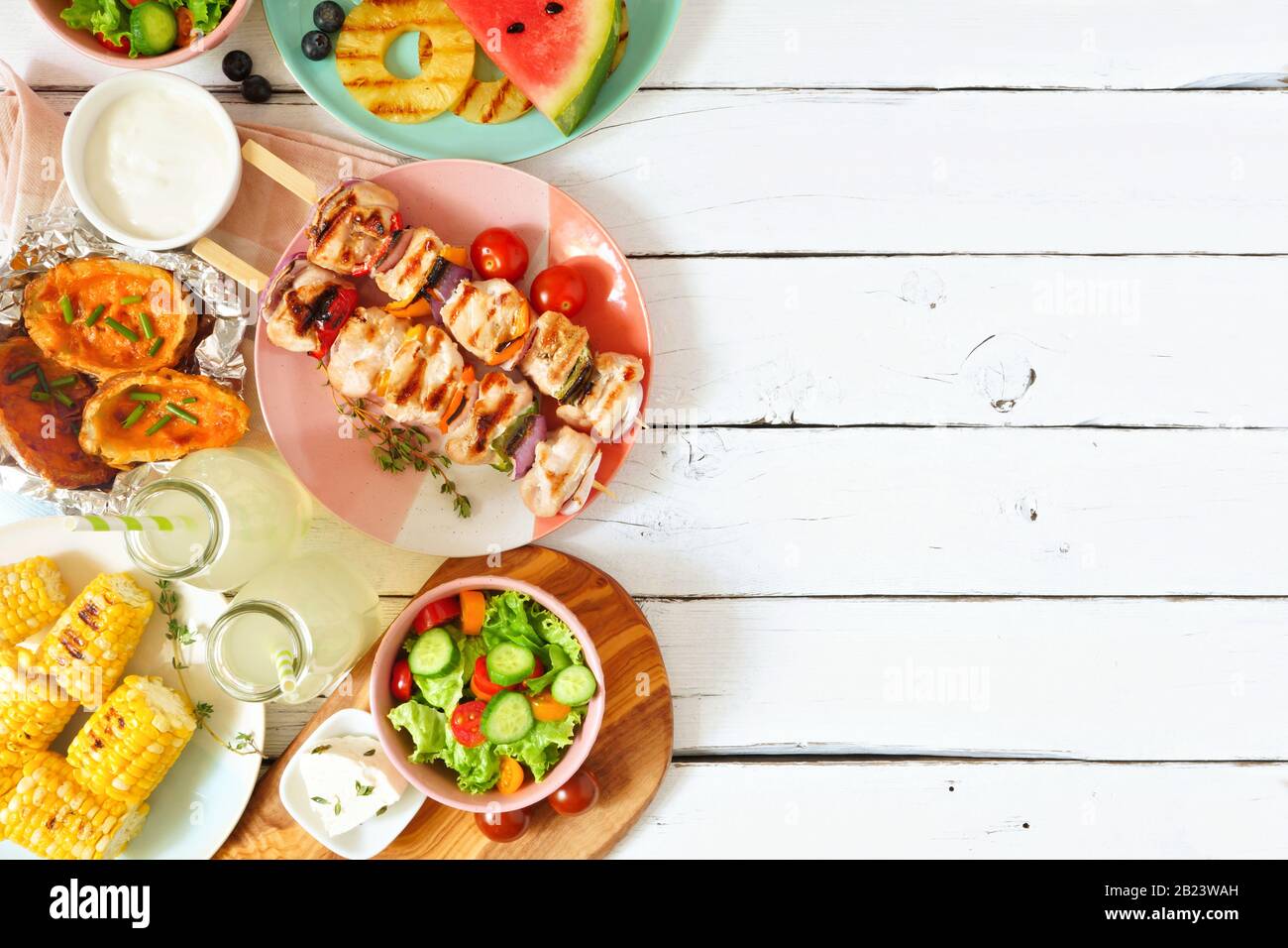 Summer BBQ or picnic food side border. Selection of grilled meat, fruits, salad and potatoes. Above view over a white wood background. Copy space. Stock Photo