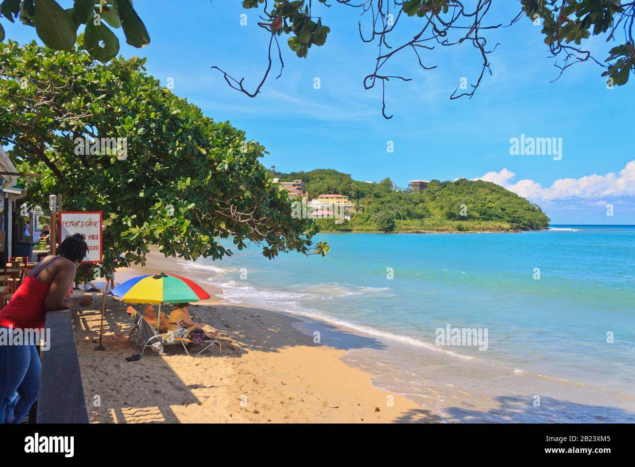 Castries, Saint Lucia - November 23, 2019. Beautiful day at Vigie tourists under multicolored beach umbrellas, local lady standing looking at them Stock Photo