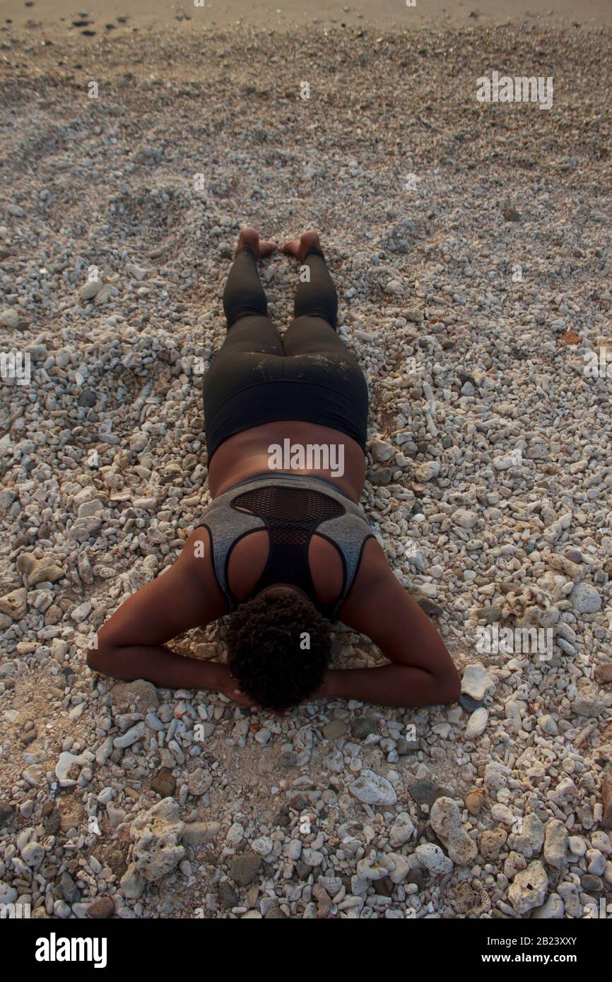 African lady the natural hair and wearing a black tights and black and gray sports bra lying face down on the coral stones and sand on the beach Stock Photo
