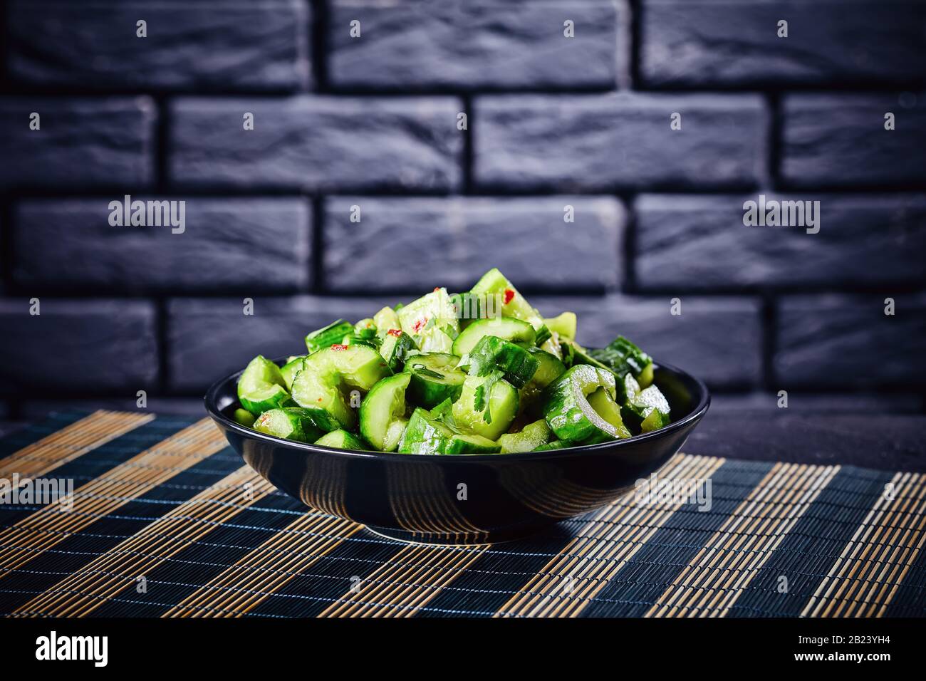 Chinese Smashed Cucumber salad, pai huang gua,  sprinkled with fresh coriander leaves in a black bowl on a concrete table with a brick wall at the bac Stock Photo