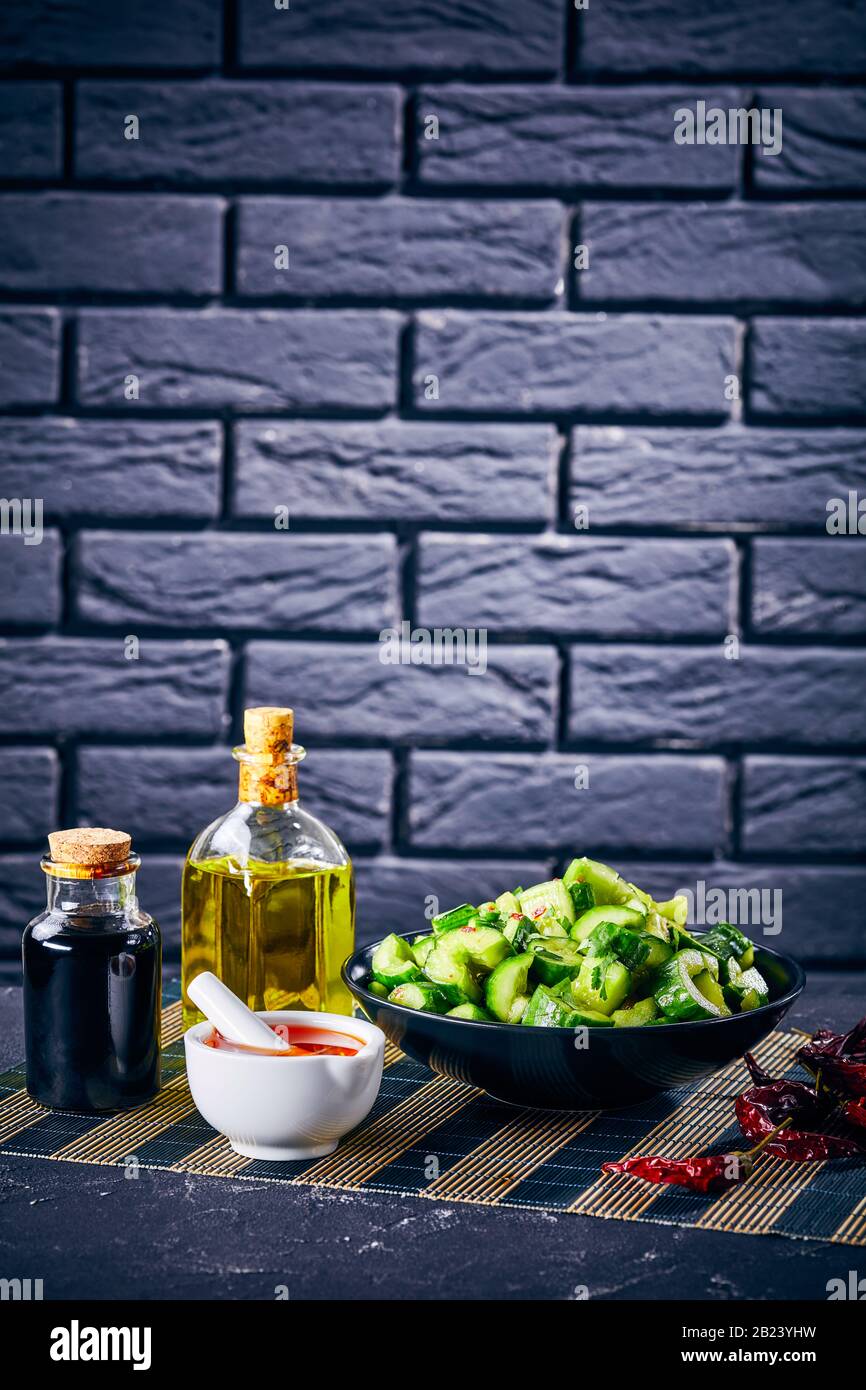 Chinese Smashed Cucumber salad, pai huang gua, in black bowl on a concrete table with ingredients and a brick wall at the background, close-up, vertic Stock Photo