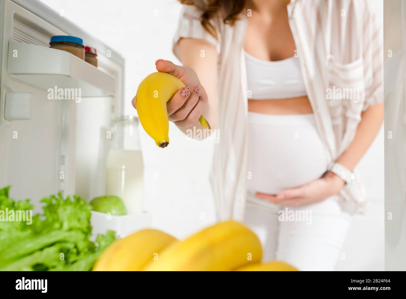 Selective focus of pregnant woman holding banana in open fridge on
