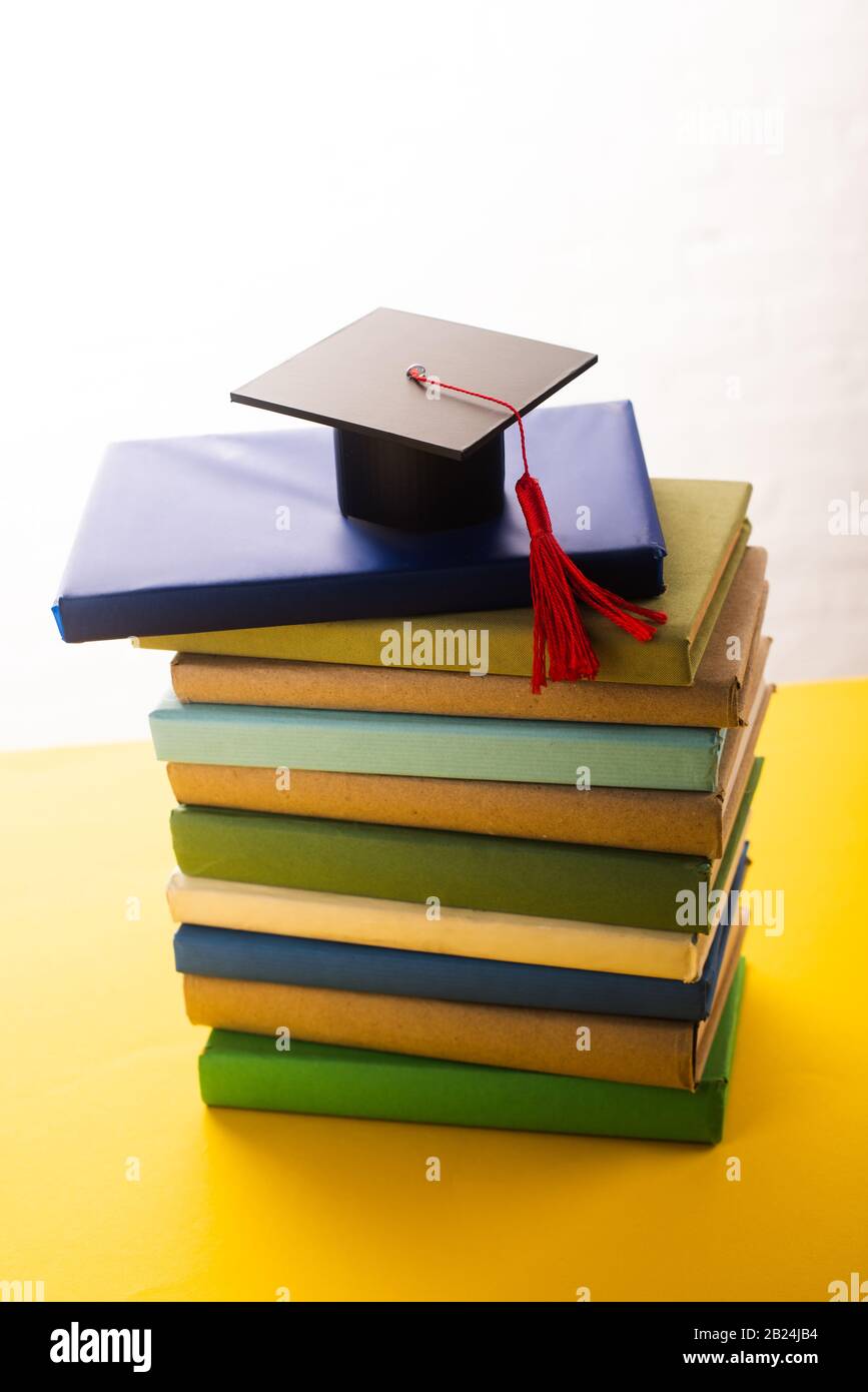 Graduation cap with red tassel on top of books on yellow surface on