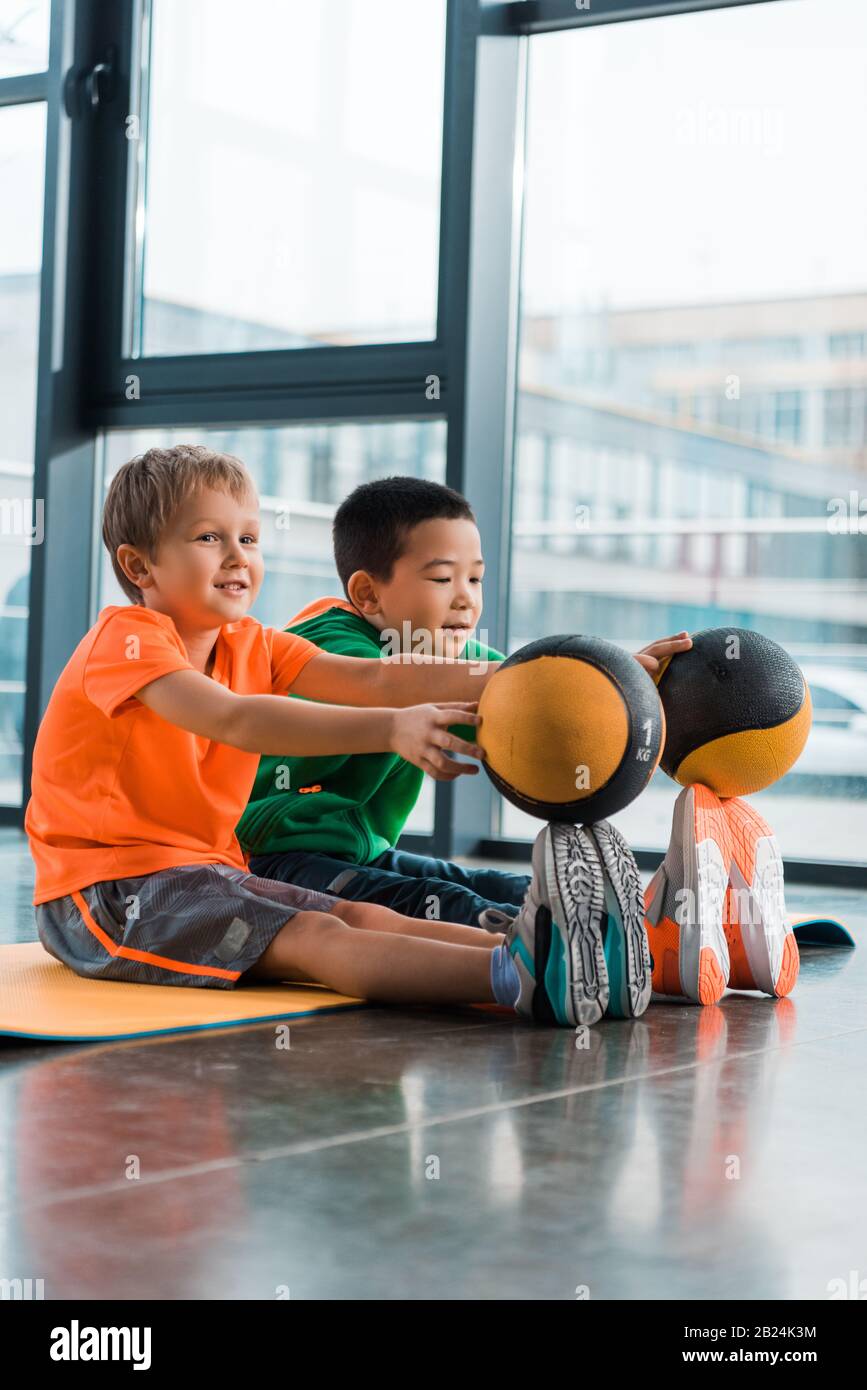 Multicultural children putting balls on tips of toes and sitting on fitness mat in gym Stock Photo