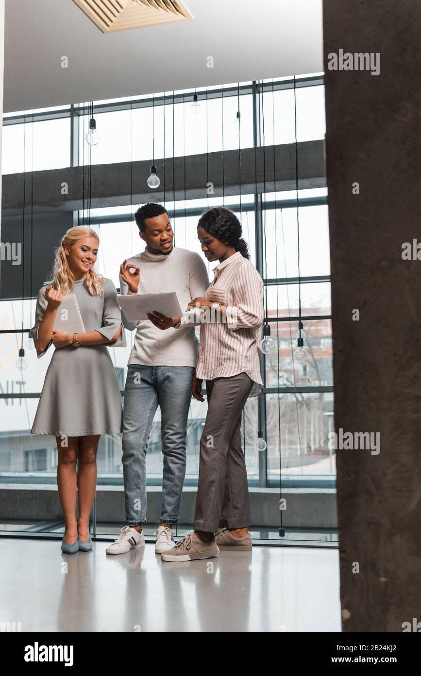 young multicultural businesspeople having discussion while standing in office and looking at documents Stock Photo