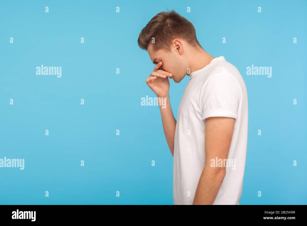 Side view of upset man in white t-shirt touching eyes, wiping away tears and crying quietly, feeling desperate and hopeless, lonely suffering depressi Stock Photo