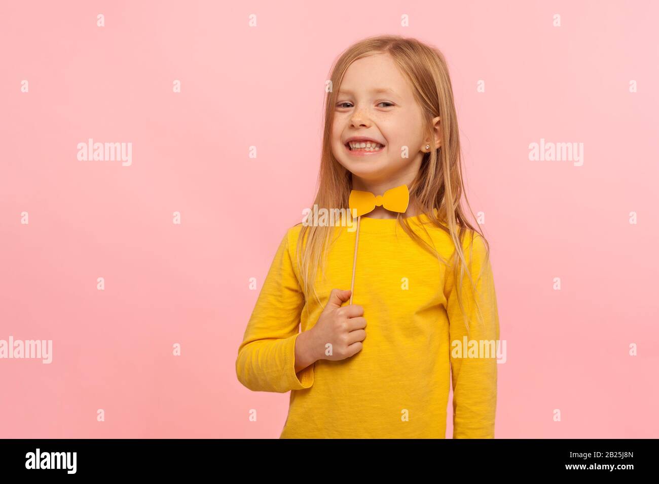 Portrait of charming playful little ginger girl holding paper bow tie and looking at camera with toothy smile, having fun, wearing masquerade accessor Stock Photo