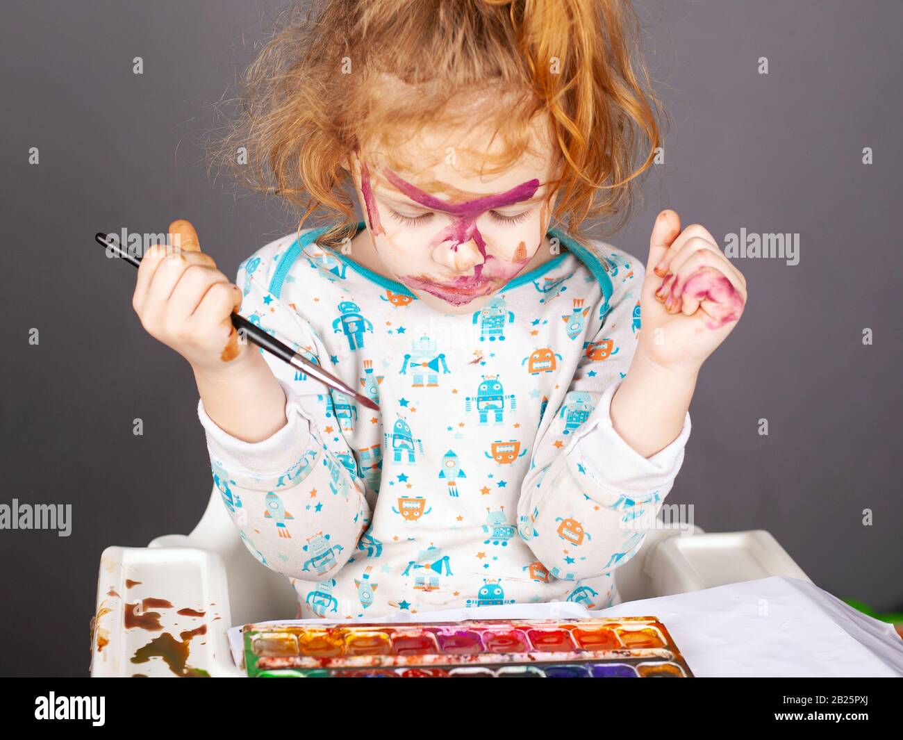 little beautiful red-haired girl is played with paints on a dark background. the child painted his face in watercolor. Stock Photo