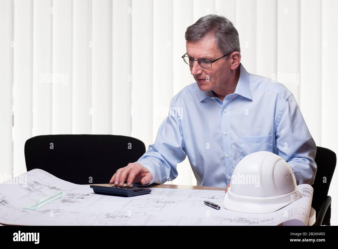 Senior architect or engineer calculating a project in an white office with blueprint, hard hat und calculator on a table Stock Photo