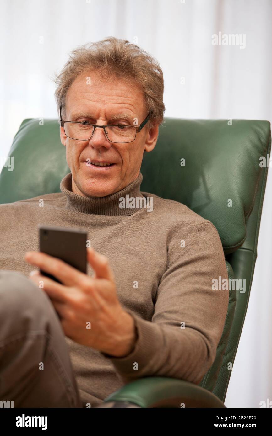 Attractive mature man with glasses looking at smartphone in a bright living room Stock Photo