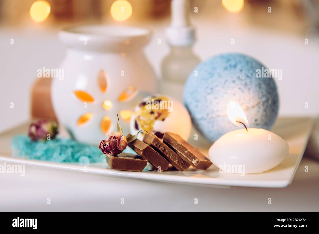 Home spa products on wooden white ceramic tray: bath bomb, aroma bath salt, aroma oil lamp, essential and massage oils, pieces of chocolate. Stock Photo