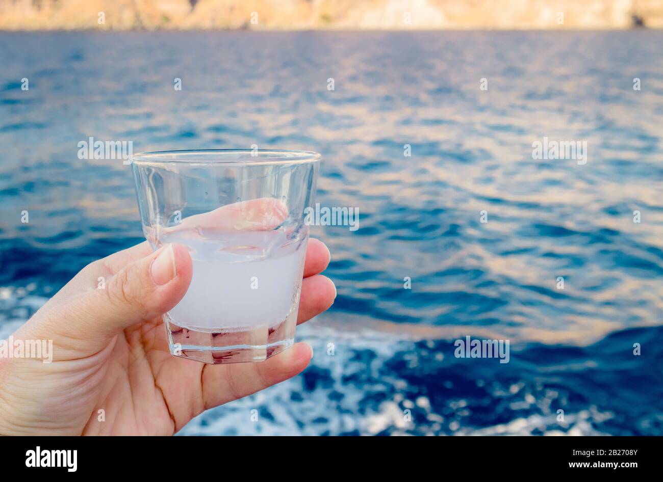 Person tourist hand holding shot glass of Ouzo witch is a dry anise-flavoured aperitif and tasting it outdoors in Santorini, Greece. Stock Photo