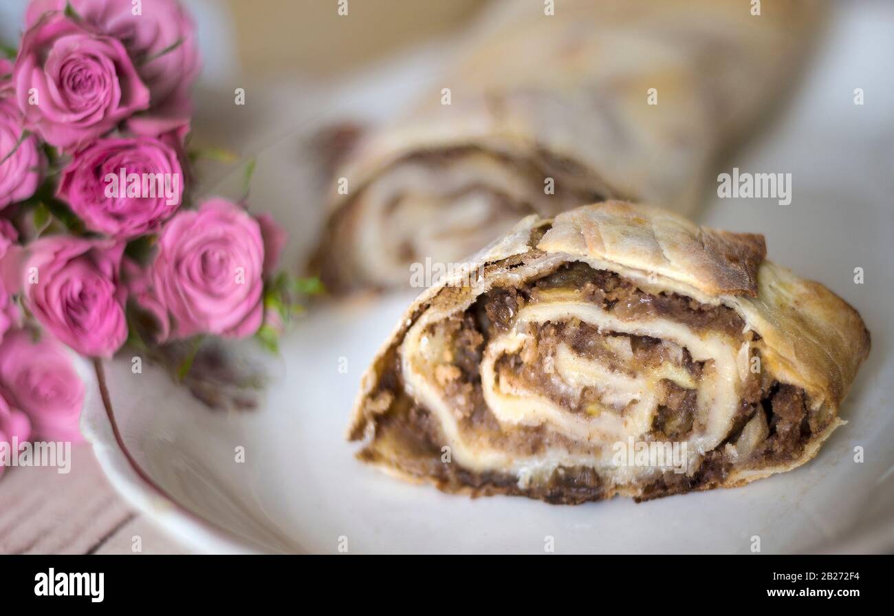 Strudel slice on a white dish. Pink roses lying nearby enliven the culinary composition. Stock Photo
