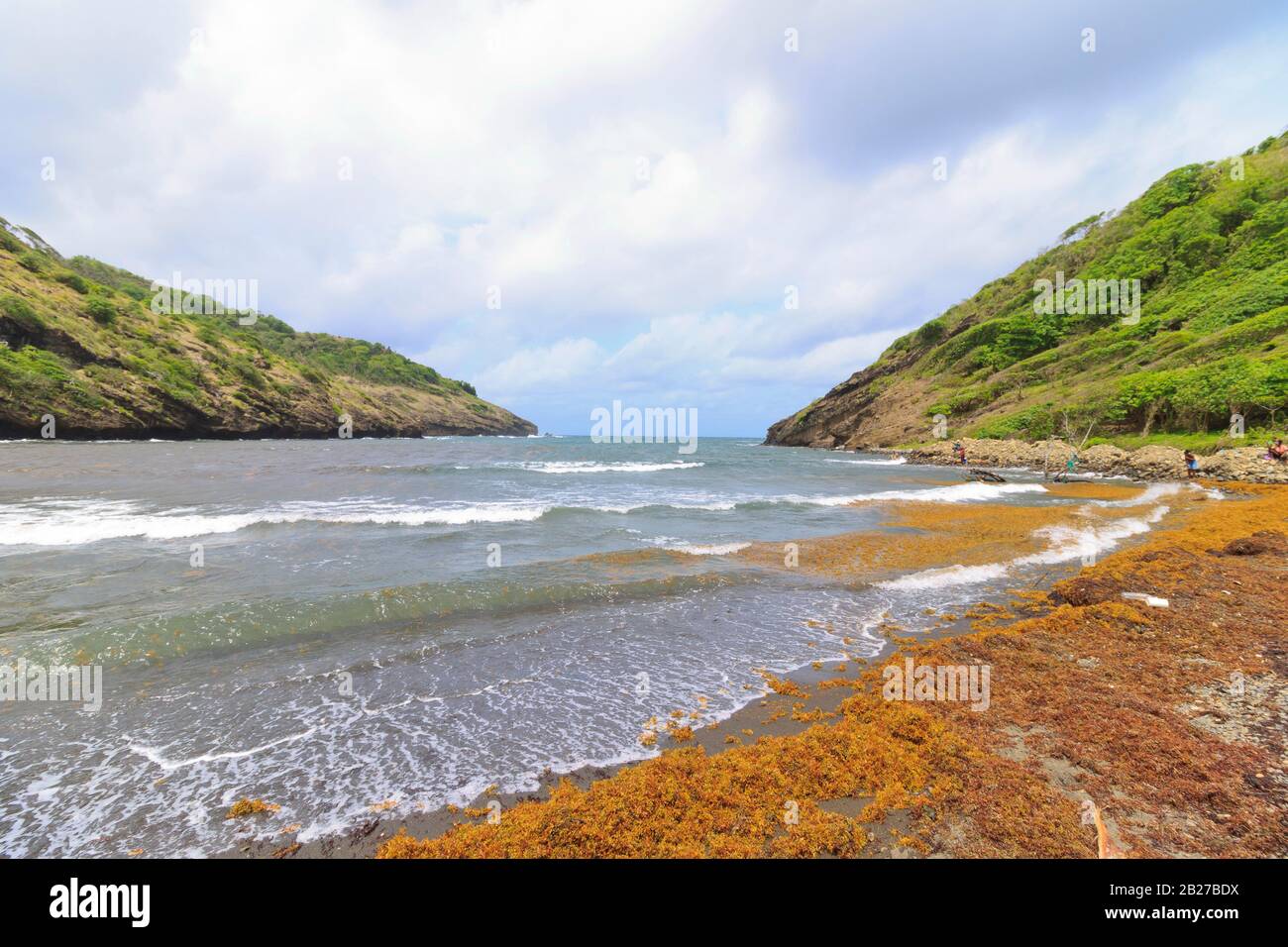 small sheltered bay in a rural village with overcast sky when the sea is not calm and black sand beach covered in seaweed with people in background Stock Photo