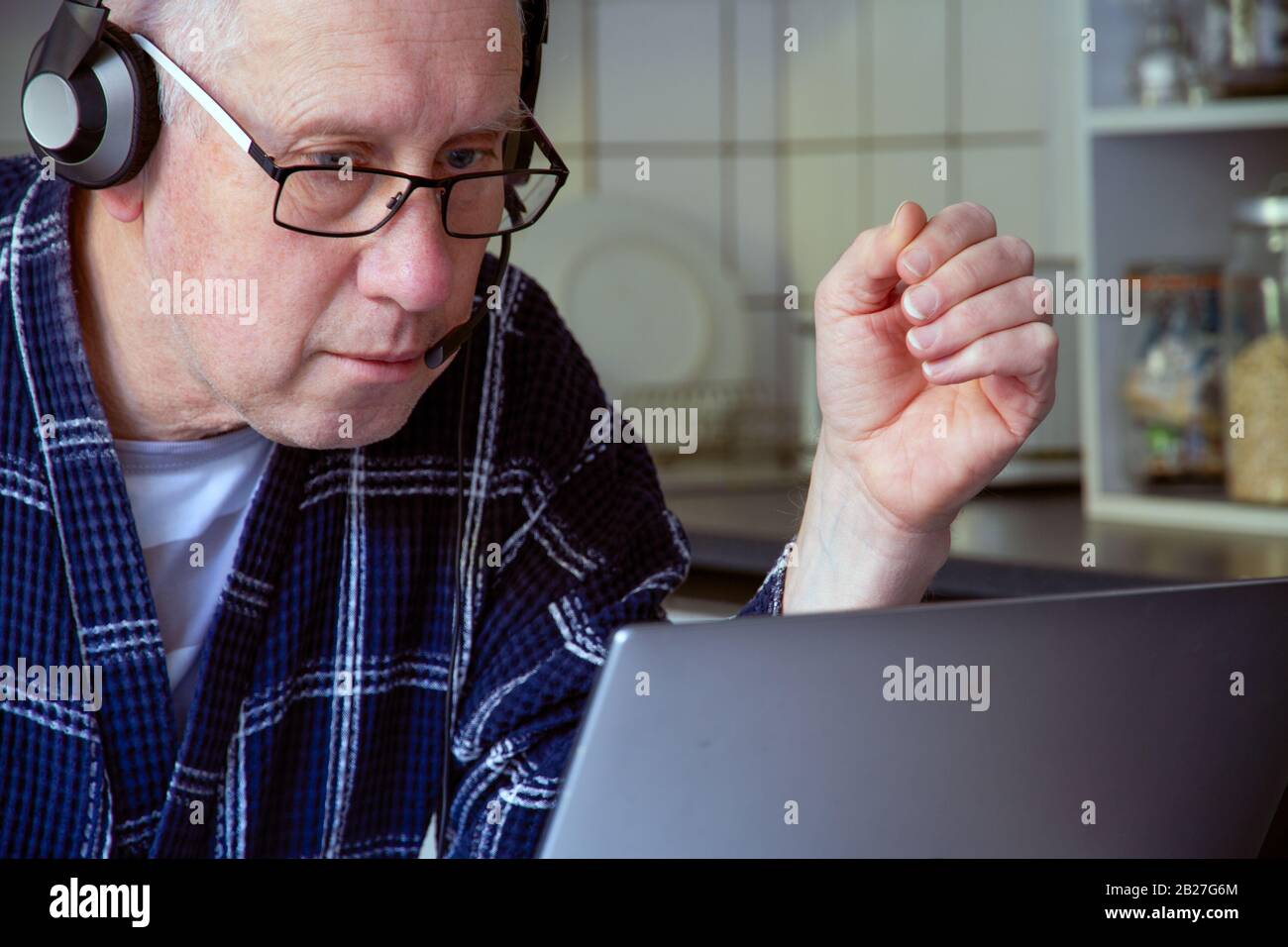 An older white man in his dressing gown and wearing headphones sits in front of a laptop in a kitchen enviromnent. Stock Photo