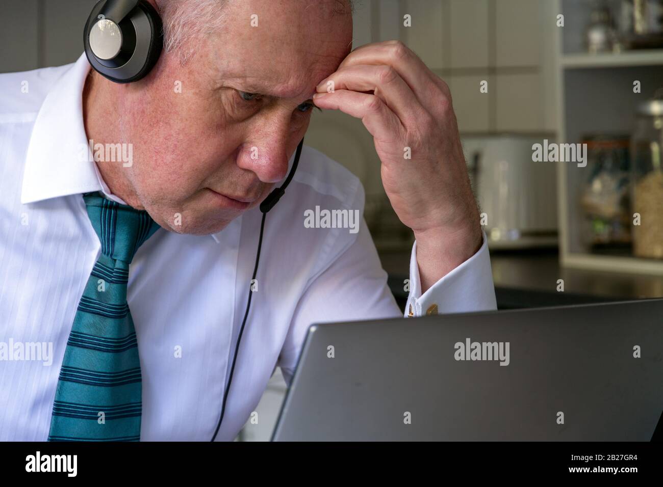 An older man in white shirt and tie wearing headphones looking frustrated and staring at laptop with evidence of working from kitchen in the backgound Stock Photo