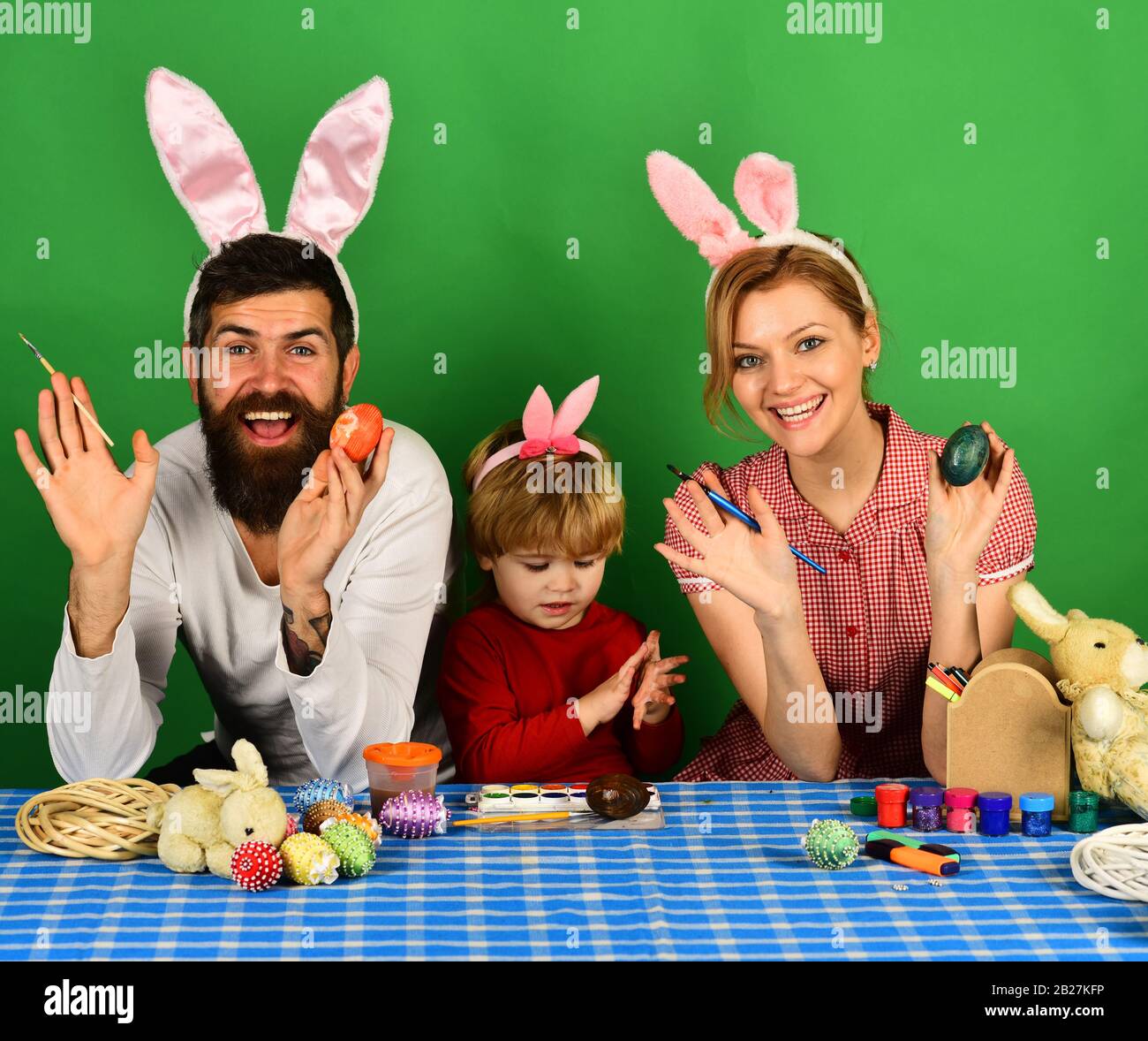 Mother, father and son painting eggs in pink bunny ears. Happy moments and Easter celebration concept. Family members preparing for Easter on green background. Man and woman with happy smiles. Stock Photo