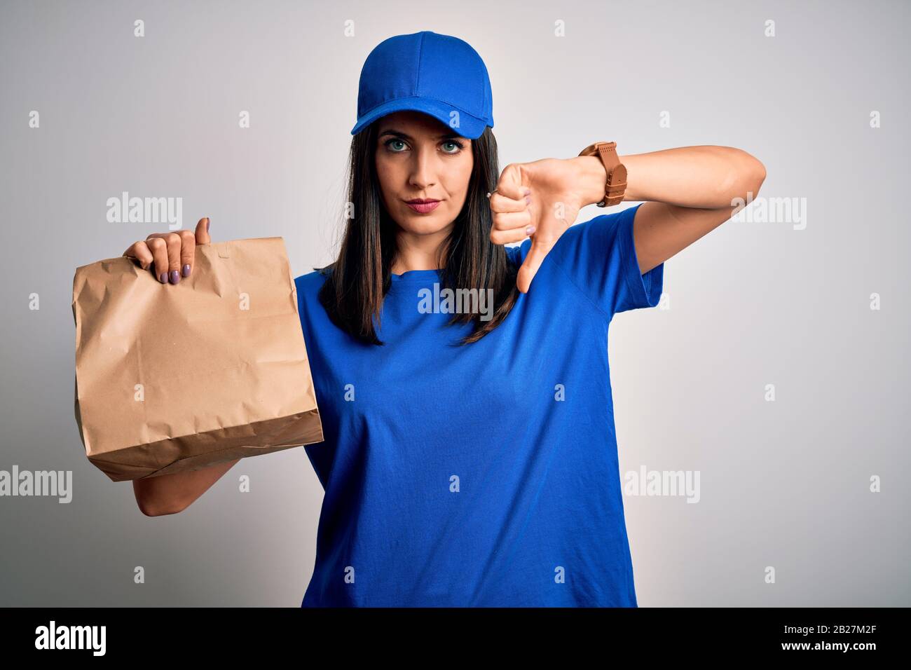 Young delivery woman with blue eyes wearing cap holding paper bag with food with angry face
