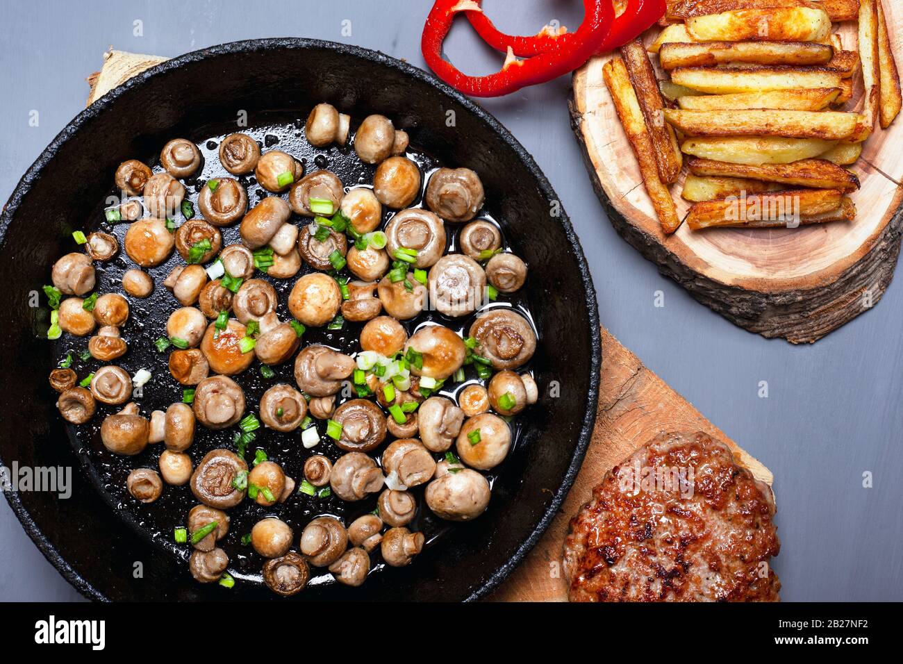 golden mushrooms champignon sprinkled with green onions in a cast iron black pan fries and juicy meat cutlet on forest wooden coasters on a gray backg Stock Photo