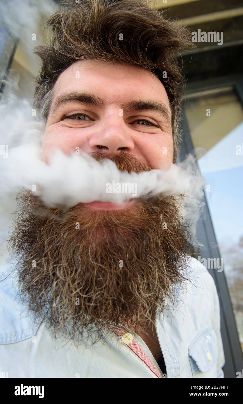 Hipster with tousled hair and gray on relaxed cheerful face with white  smoke flying out of mouth. Vaping concept. Man with beard and mustache  smoking, building background Stock Photo