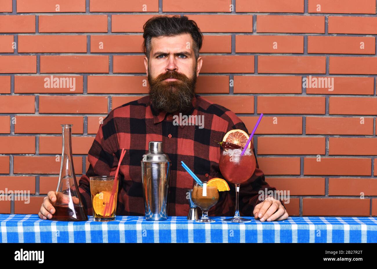 Man in checkered shirt on brick wall background prepares drinks. Barman with long beard and mustache and stylish hair on serious face made alcoholic cocktails. Alcoholic drinks concept Stock Photo