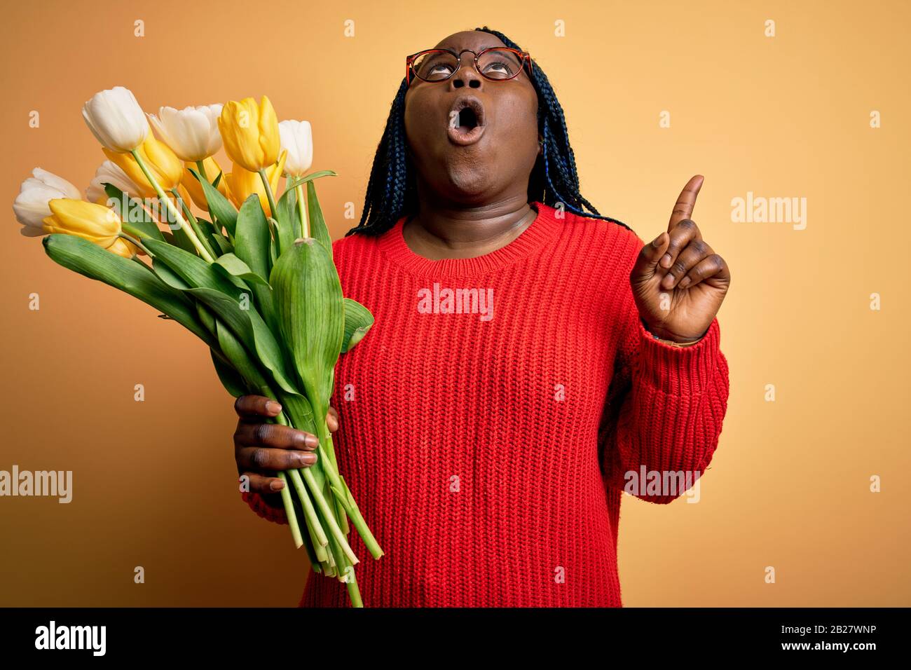 Young african american plus size woman with braids holding bouquet of yellow tulips flower amazed and surprised looking up and pointing with fingers a Stock Photo