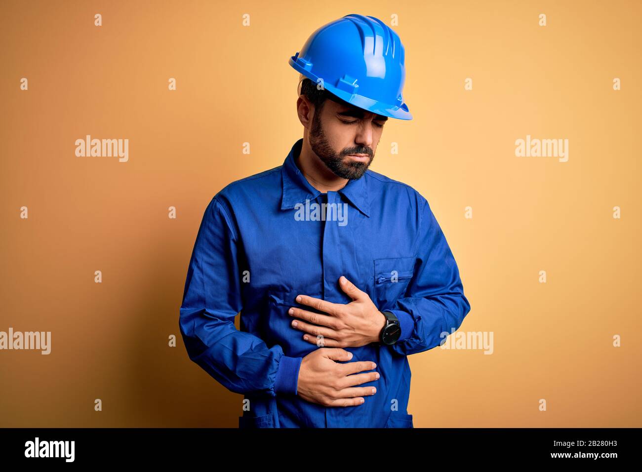 Mechanic man with beard wearing blue uniform and safety helmet over yellow background with hand on stomach because indigestion, painful illness feelin Stock Photo