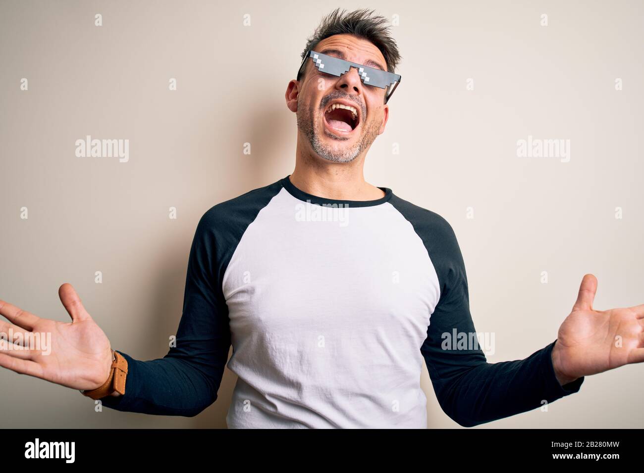 Young handsome man wearing funny thug life sunglasses meme over white background crazy and mad shouting and yelling with aggressive expression and arm Stock Photo
