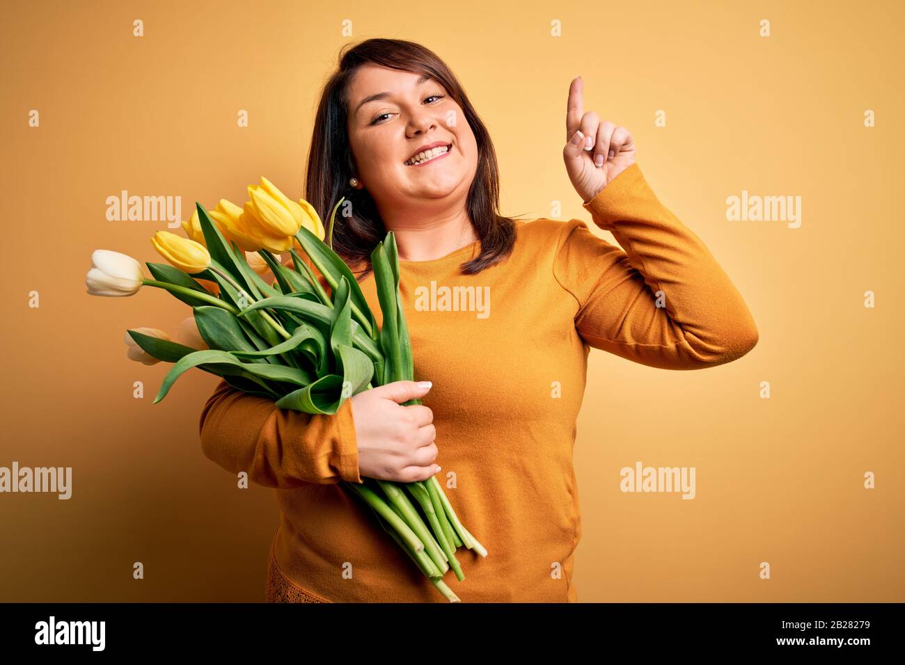 Beautiful plus size woman holding romantic bouquet of natural tulips flowers over yellow background smiling amazed and surprised and pointing up with Stock Photo