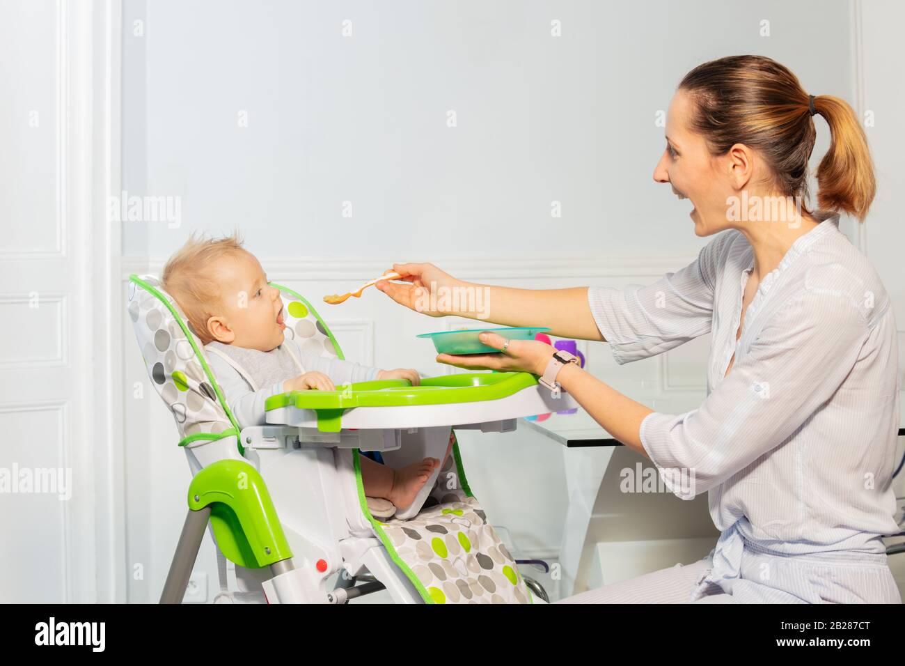 Happy little toddler baby boy sit in highchair and eat from plastic spoon with mother help in mouse looking at camera Stock Photo